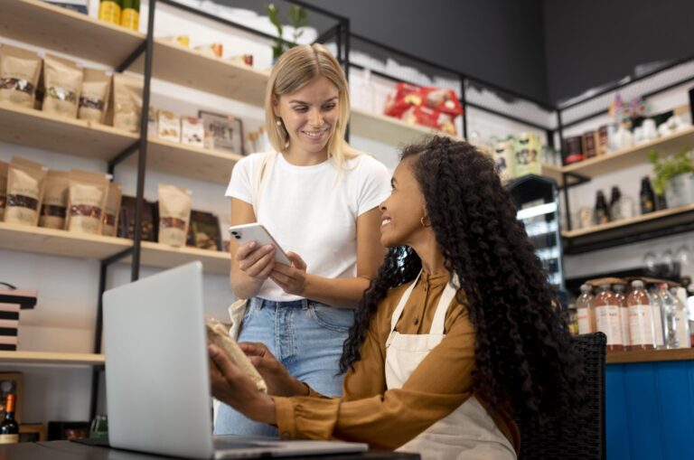 Empreendedora de avental sentada ao notebook conversa com cliente que mostra o celular, em loja com prateleiras de produtos embalados